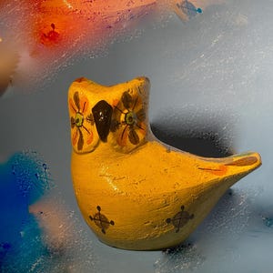 May include: A yellow ceramic owl figurine with painted details, including black eyes, brown beak, and floral patterns. The owl has a textured surface and is sitting on a light blue background.