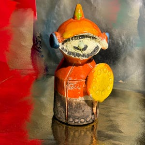 May include: A whimsical ceramic figurine in shades of orange, yellow, and black. The figure has a helmet-like head with blue accents, a smiling face, and a round yellow shield. The base has a textured, dark finish.