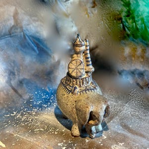 Pode incluir: Estatueta de elefante de cerâmica com um design detalhado. O elefante é marrom claro com detalhes em azul escuro e uma figura nas costas. A estatueta tem uma superfície texturizada e uma aparência artística única.