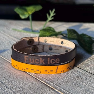 May include: A leather bracelet with the text "Fuck Ice" engraved on it. The bracelet is black and yellow, with a Morse code pattern on the yellow band. The bracelet is secured with two metal snaps.
