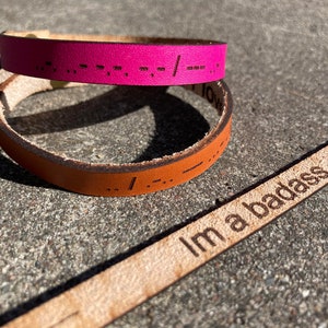 May include: Two leather bracelets with braille lettering. The top bracelet is pink and the bottom bracelet is brown. A third leather strap is laying on top of the bracelets with the text "I'm a badass" engraved on it.