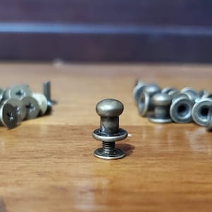 May include: A collection of antique brass-colored screws and rivets are arranged on a wooden surface. The screws have flat heads with cross-shaped slots, while the rivets have a rounded top and a flat base. The hardware is likely for furniture or craft projects.