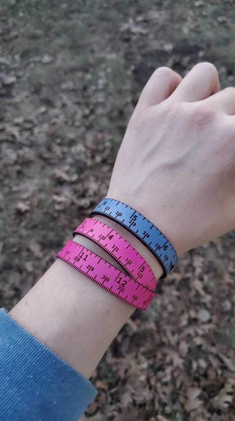 May include: Three fabric measuring tape bracelets. The top bracelet is blue, the middle bracelet is pink, and the bottom bracelet is pink. The bracelets are stacked on top of each other on a person's wrist.