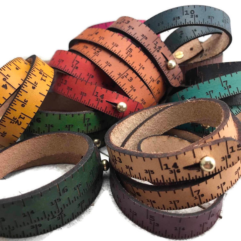 May include: A collection of leather bracelets with ruler markings. The bracelets are in various colors, including green, red, brown, and black. Each bracelet has a brass rivet.
