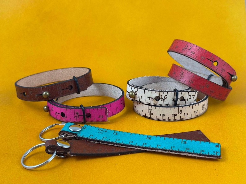 May include: Four leather bracelets with a measuring tape design. The bracelets are in pink, red, and white. There is also a brown leather keychain with a blue measuring tape.