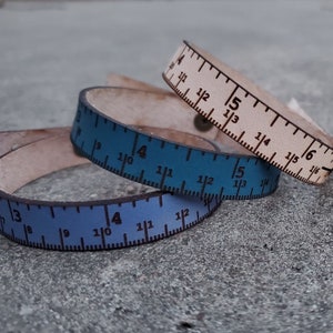 May include: Three leather bracelets with ruler markings. The bracelets are in shades of blue and white. The markings on the bracelets are in inches.