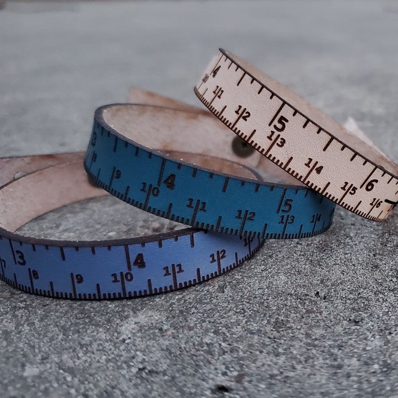 May include: Three leather bracelets with ruler markings. The bracelets are in shades of blue and white. The markings on the bracelets are in inches.