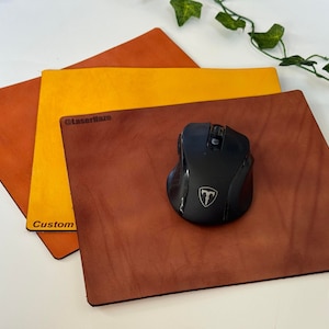 May include: Three leather mouse pads in different shades of brown and yellow. The top pad is yellow, the middle pad is brown, and the bottom pad is a darker shade of brown. The word "Custom" is engraved on the middle pad, and the word "@Laserilaz" is engraved on the top pad. A black computer mouse is on top of the bottom pad.