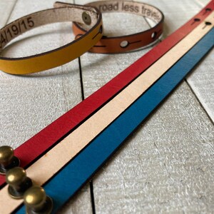 May include: Three leather bracelets with different colors and designs. The first bracelet is yellow and has the date "04/19/15" engraved on it. The second bracelet is brown and has the text "road less traveled" engraved on it. The third bracelet is multi-colored with red, white, and blue stripes and has brass rivets.
