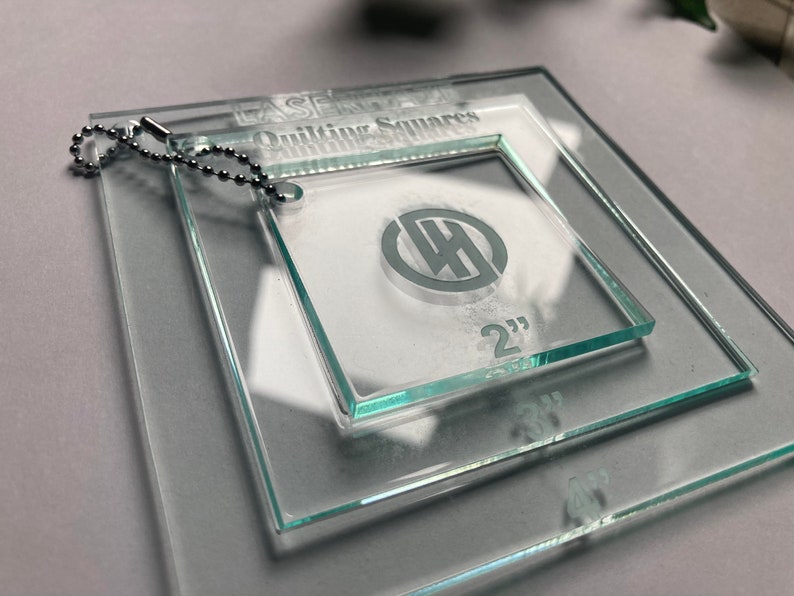 May include: Set of three clear glass quilting squares with a keychain. The squares are 2 inches, 3 inches, and 4 inches. The squares have a logo with the letters "WH" inside a circle.