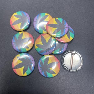 May include: Ten rainbow-colored button pins with a black marijuana leaf design. The pins are arranged in a circle with one pin in the center.