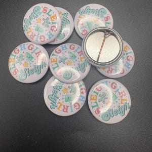 May include: A collection of white round buttons with the text "Sleigh Girl" in a colorful, retro font. Each button features a gingerbread man and a Christmas tree design. One button has a pin back.