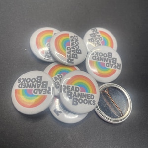 May include: A close-up of several rainbow-colored button pins with the text "Read Banned Books" in black. The buttons are scattered on a dark gray surface.