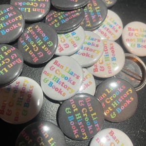 May include: A collection of round buttons with the text "Ban Liars & Crooks not History & Books" in rainbow colors. The buttons are in various shades of gray and white.