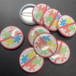 Pu&ograve; includere: Un set di otto spille rotonde con un cerchio rosso attorno a uno sfondo bianco. Lo sfondo bianco ha una linea rossa che lo attraversa e un design a puzzle multicolore. I pezzi del puzzle sono blu, gialli e verdi.