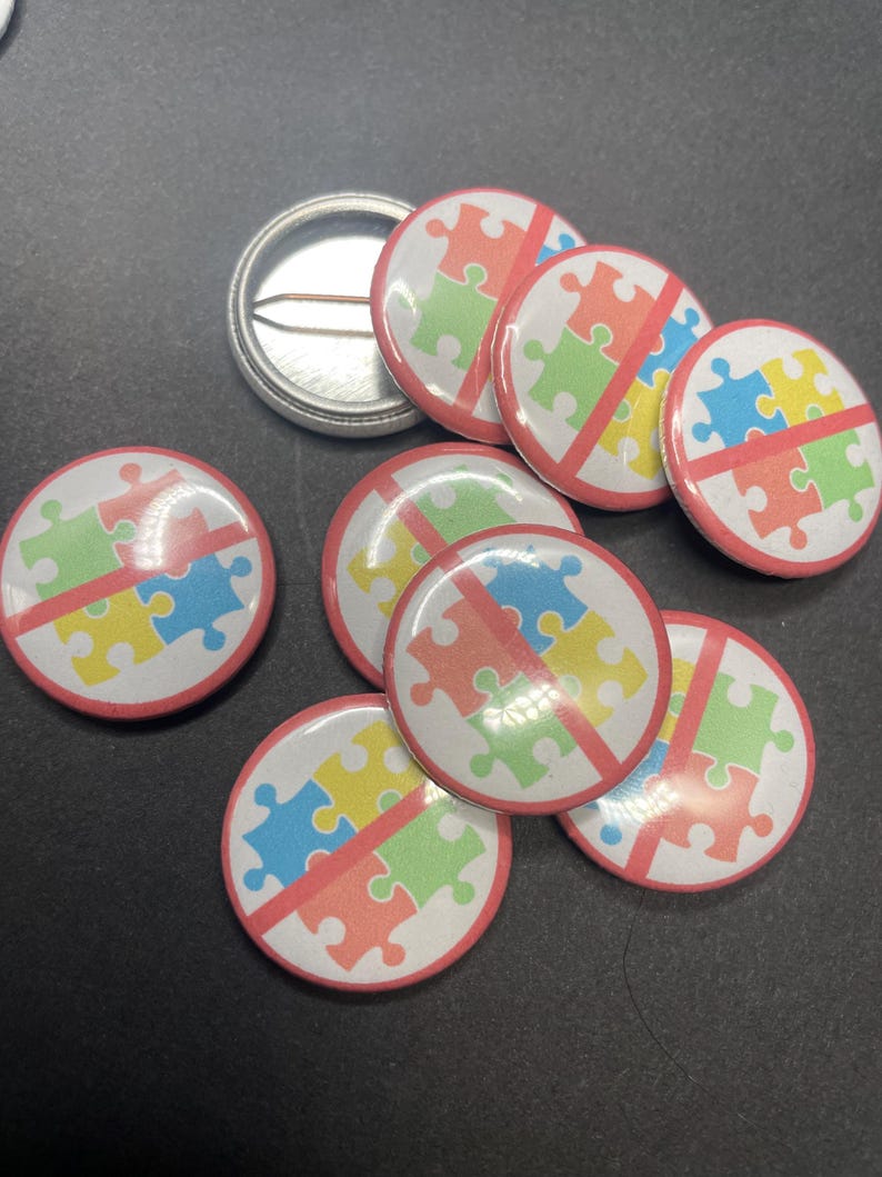 Pu&ograve; includere: Un set di otto spille rotonde con un cerchio rosso attorno a uno sfondo bianco. Lo sfondo bianco ha una linea rossa che lo attraversa e un design a puzzle multicolore. I pezzi del puzzle sono blu, gialli e verdi.