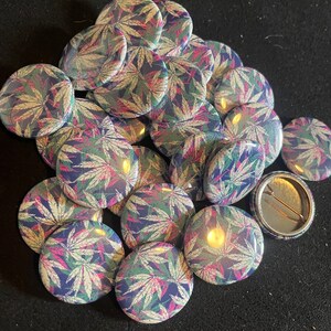 May include: A pile of round, colorful buttons with a marijuana leaf design. The buttons are about 1 inch in diameter and have a silver metal backing.