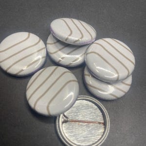 May include: Six white round buttons with a brown striped pattern. The buttons have a silver back with a pin.