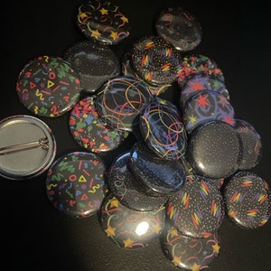May include: A collection of 17 round black button pins with various colorful patterns, including confetti, stars, and swirls.