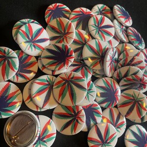 May include: A collection of round, white button pins with a green, red, and blue marijuana leaf design.
