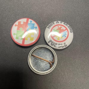 Pu&ograve; includere: Tre bottoni rotondi, due con una barra rossa che attraversa un pezzo di puzzle multicolore. Il bottone superiore dice "NOT A F*CKING PUZZLE". Il bottone inferiore &egrave; argentato con uno spillo sul retro.
