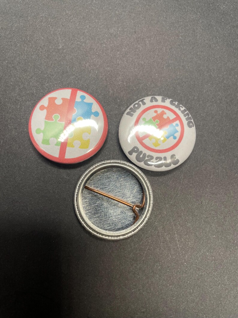 Pu&ograve; includere: Tre bottoni rotondi, due con una barra rossa che attraversa un pezzo di puzzle multicolore. Il bottone superiore dice "NOT A F*CKING PUZZLE". Il bottone inferiore &egrave; argentato con uno spillo sul retro.