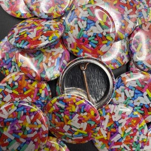 May include: A collection of colorful button pins with a glossy finish. Each pin features a vibrant sprinkle design in shades of red, yellow, blue, green, and white. The pins have a metal backing with a safety pin closure.