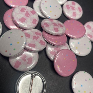 May include: A collection of pink and white button pins with various designs. Some buttons feature a pink background with white sprinkles, while others have a white background with colorful sprinkles. A few buttons have a pink background with a white cow print design.