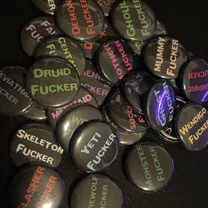 May include: A collection of black and white button pins with various mythical creatures and the word "Fucker" written in colorful dripping paint. The pins include a dragon, a ghoul, a mummy, a werewolf, a yeti, a skeleton, a slasher, a druid, a mermaid, a succubus, a wendigo, a demon, a centaur, a ghost, a crypt, a leviathan, and a monster.