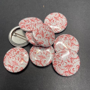 May include: A group of eight round white button pins with a pink splatter paint design.