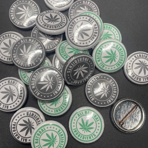 May include: A collection of round, white button pins with a green marijuana leaf design. The pins have the text "All Natural Ingredients" printed around the leaf.