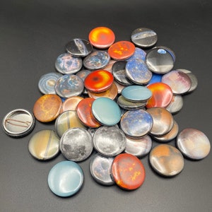 May include: A collection of colorful round buttons with space-themed designs, including planets, stars, and galaxies. The buttons are arranged in a pile on a black surface.