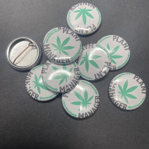 May include: A group of eight white round buttons with a green marijuana leaf design and the text "Plant Manager" in black.