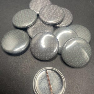 May include: A close-up of a pile of round, black and silver buttons with a white binary code pattern. The buttons are arranged in a random pattern. One button is lying on its back, showing the pin on the back.