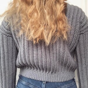 May include: A gray knitted sweater with a ribbed texture. The sweater is cropped and has long sleeves.