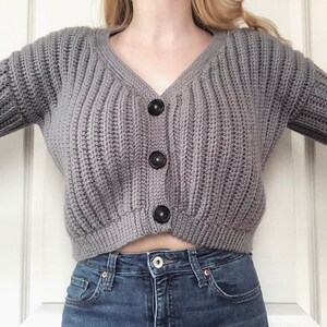 May include: A gray knitted cardigan with a v-neck and black buttons. The cardigan is cropped and has a ribbed texture.