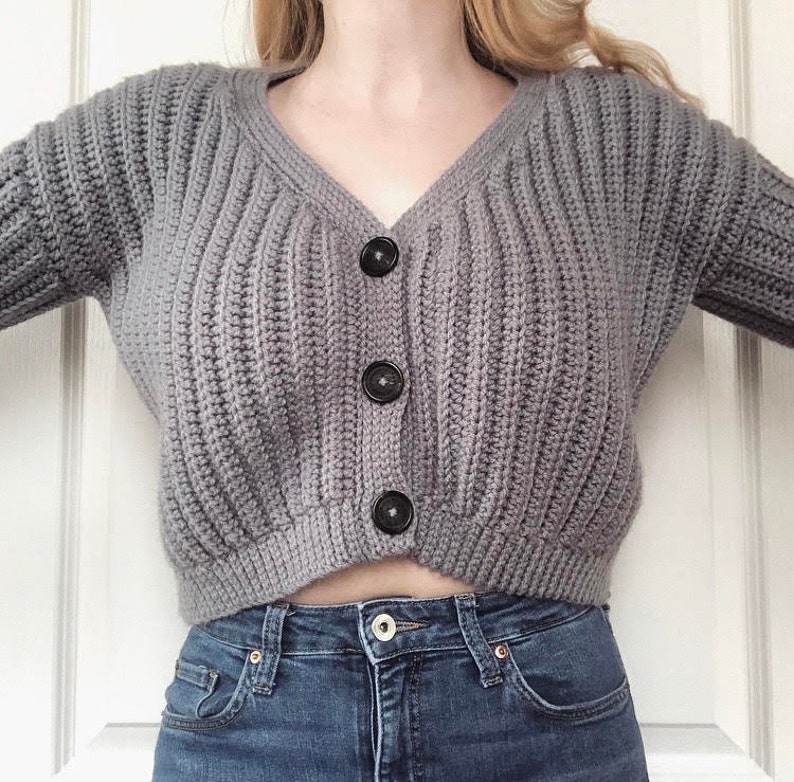 May include: A gray knitted cardigan with a v-neck and black buttons. The cardigan is cropped and has a ribbed texture.