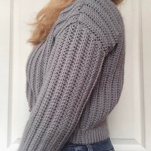 May include: A gray crocheted sweater with a ribbed texture. The sweater has long sleeves and a cropped length.