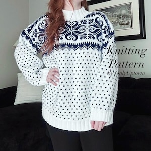 May include: A white and navy blue knitted sweater with a snowflake design on the upper chest and sleeves. The sweater has a high, ribbed collar and is covered in small navy blue heart shapes. The text "Knitting Pattern By Only Uptown" is visible.