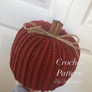 May include: Crocheted pumpkin with a cinnamon stick stem and twine bow. The pumpkin is a deep orange colour and has a ribbed texture. The text "Crochet Pattern By Only Uptown" is visible at the bottom of the image.