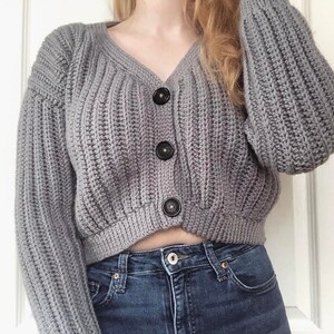 May include: A gray knitted cardigan with a v-neck and three black buttons. The cardigan is cropped and has long sleeves.