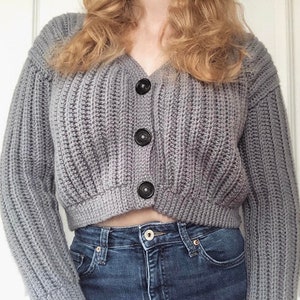 May include: A gray cropped cardigan with a v-neck and four black buttons. The cardigan is made of a soft, knitted fabric.