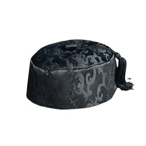 Puede incluir: Un fez negro de forma cilíndrica con la parte superior plana y una borla negra a juego sujeta en el centro. La tela presenta un sutil patrón jacquard tonal con motivos orgánicos y ondulados en toda la superficie del sombrero.