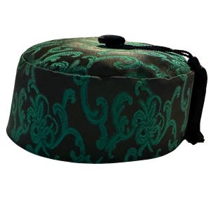 Puede incluir: Un gorro fez negro con un intrincado diseño floral verde. El sombrero tiene una parte superior redondeada, una base cilíndrica y una borla negra. La tela tiene un ligero brillo y el diseño es simétrico.