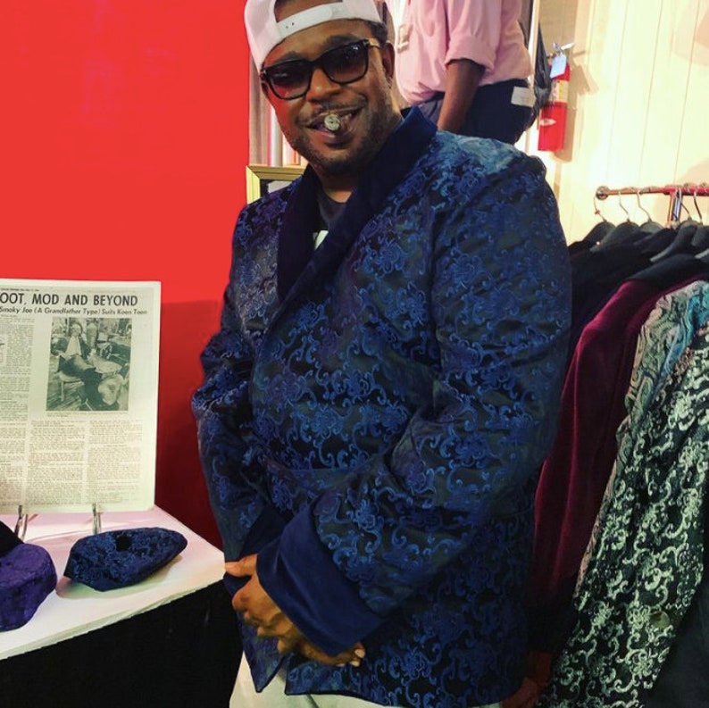 May include: A man wearing a blue patterned blazer with a velvet collar and a white hat. He is holding a cigar in his mouth and looking at the camera. The blazer has a floral pattern and the man is standing in front of a red wall.