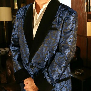 May include: A man wearing a blue and black patterned silk robe with a black velvet collar. The robe has a shawl collar and is open in the front. The man is standing in a room with a wooden bookcase and a window in the background.