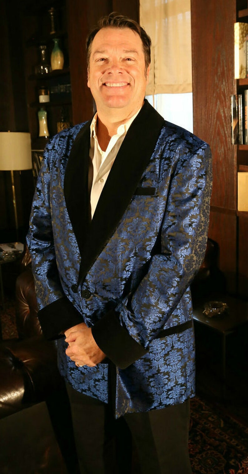 May include: A man wearing a blue and black patterned silk robe with a black velvet collar. The robe has a shawl collar and is open in the front. The man is standing in a room with a wooden bookcase and a window in the background.