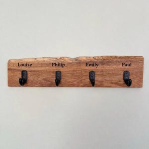 May include: A rectangular wooden wall hook rack with a natural live edge top. The board features four black metal hooks and the engraved names Louise, Philip, Emily, and Paul centered above each hook in a dark, clear serif font.