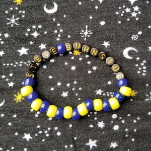 May include: A blue and yellow beaded bracelet with the words "Steve Burnside" spelled out in black letters.