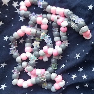May include: A multi-colored kandi bracelet with pink, white, and gray beads and iridescent star-shaped charms.